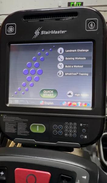 A close-up of a StairMaster treadmill console showing the touch screen interface with options like Landmark Challenge, Existing Workouts, and Quick Start. The black console has a green power button and a keypad below the screen.